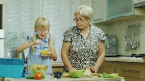 Ein 6-jähriges Mädchen in den rosa Gläsern bereitet einen Salat in der Küche zu Ihre Großmutterhilfen, benutzen eine Tablette stock video footage Ein 6-jähriges Mädchen in den rosa Gläsern bereitet einen Salat in der Küche zu Ihre Großmutterhilfen, benutzen eine Tablette stock video footage