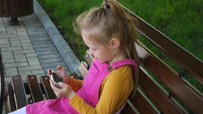 Ein lächelndes Mädchen ein Vorschulalter, benutzt das Telefon draußen im Park Sonniger Sommertag stock video Ein lächelndes Mädchen ein Vorschulalter, benutzt das Telefon draußen im Park Sonniger Sommertag stock video