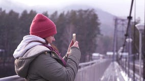 Ein Mädchen, das auf einer Brücke in einer Jacke steht, macht ein Panorama von der Landschaft an ihrem Telefon Zeitlupe, 1920x108 stock video Ein Mädchen, das auf einer Brücke in einer Jacke steht, macht ein Panorama von der Landschaft an ihrem Telefon Zeitlupe, 1920x108 stock video