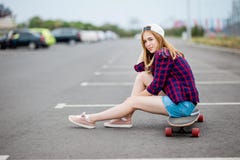 Ein schönes blondes Mädchen, das karierte Hemd-, Kappen- und Denimkurze hosen trägt, sitzt auf dem longboard auf dem Parkplatz mi lizenzfreie stockfotografie Ein schönes blondes Mädchen, das karierte Hemd-, Kappen- und Denimkurze hosen trägt, sitzt auf dem longboard auf dem Parkplatz mi lizenzfreie stockfotografie