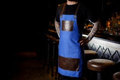 El camarero joven con el tatuaje en las manos se vistió en delantal azul y marrón imagen de archivo El camarero joven con el tatuaje en las manos se vistió en delantal azul y marrón imagen de archivo