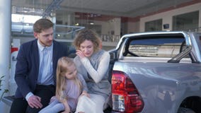 El centro de venta auto, pareja joven con el niño elige el automóvil y comunica con uno a mientras que se sienta en tronco en almacen de metraje de vídeo El centro de venta auto, pareja joven con el niño elige el automóvil y comunica con uno a mientras que se sienta en tronco en almacen de metraje de vídeo