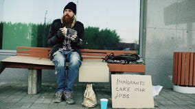 El hombre europeo sin hogar y desempleado con la muestra de la cartulina come el bocadillo en banco en la calle de la ciudad debi fotos de archivo libres de regalías El hombre europeo sin hogar y desempleado con la muestra de la cartulina come el bocadillo en banco en la calle de la ciudad debi fotos de archivo libres de regalías