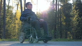 El hombre físicamente desafiado se está sentando en su silla de ruedas accionada al lado del bosque almacen de metraje de vídeo El hombre físicamente desafiado se está sentando en su silla de ruedas accionada al lado del bosque almacen de metraje de vídeo