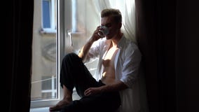 El hombre joven hermoso y atractivo con el torso desnudo bebe el café que se sienta en el alféizar por la mañana metrajes El hombre joven hermoso y atractivo con el torso desnudo bebe el café que se sienta en el alféizar por la mañana metrajes