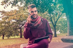 El hombre joven trabaja en la tableta mientras que habla en el teléfono al aire libre en espacio público cerca de parque imagen de archivo El hombre joven trabaja en la tableta mientras que habla en el teléfono al aire libre en espacio público cerca de parque imagen de archivo