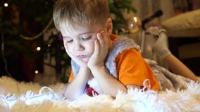 El niño miente en una manta blanca suave en el cuarto de niños Él está mirando historietas en el smartphone Navidad almacen de video El niño miente en una manta blanca suave en el cuarto de niños Él está mirando historietas en el smartphone Navidad almacen de video