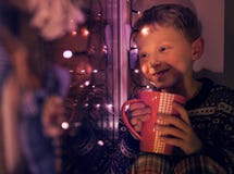 El niño pequeño con la taza grande de bebida caliente mira en la ventana con las luces de Christmass imágenes de archivo libres de regalías El niño pequeño con la taza grande de bebida caliente mira en la ventana con las luces de Christmass imágenes de archivo libres de regalías