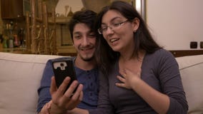 El par afectivo feliz que se sienta en el vídeo del sofá en casa que charla con los amigos parents usando el teléfono elegante almacen de metraje de vídeo El par afectivo feliz que se sienta en el vídeo del sofá en casa que charla con los amigos parents usando el teléfono elegante almacen de metraje de vídeo