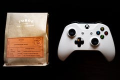 El regulador de los juegos del Xbox One se sentó al lado de un bolso del café molido contra un fondo negro oscuro imágenes de archivo libres de regalías El regulador de los juegos del Xbox One se sentó al lado de un bolso del café molido contra un fondo negro oscuro imágenes de archivo libres de regalías