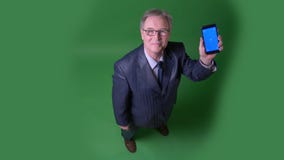 el tiro superior de la Lleno-altura del hombre mayor da vuelta en el app y muestra la pantalla azul del teléfono en fondo verde d almacen de video el tiro superior de la Lleno-altura del hombre mayor da vuelta en el app y muestra la pantalla azul del teléfono en fondo verde d almacen de video