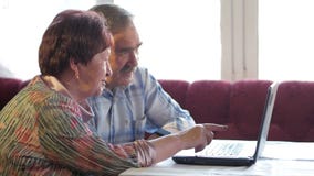 An elderly couple is sitting at home at the laptop. A woman reads news, a man with a mustache sits next to him and talks stock footage An elderly couple is sitting at home at the laptop. A woman reads news, a man with a mustache sits next to him and talks stock footage
