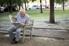 An elderly gray-haired man carefully reads a newspaper while sitting on a bench in the park stock photos An elderly gray-haired man carefully reads a newspaper while sitting on a bench in the park stock photos