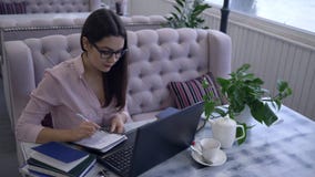 ELearning technologia, uśmiechnięta dziewczyna z piórem pisze notatkach w notatnika obsiadaniu przy stołem z laptopem w kawiarni zdjęcie wideo ELearning technologia, uśmiechnięta dziewczyna z piórem pisze notatkach w notatnika obsiadaniu przy stołem z laptopem w kawiarni zdjęcie wideo