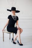 Elegant kvinna i svart klänning med ett hattsammanträde på stol arkivfoton Elegant kvinna i svart klänning med ett hattsammanträde på stol arkivfoton