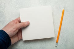An empty square piece of paper in a man`s hand, a yellow pencil next to it. Mock-up stock image An empty square piece of paper in a man`s hand, a yellow pencil next to it. Mock-up stock image