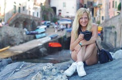 En blond flicka med ett hjärtaarmband sitter på vaggar och rymmer en kamera på Riomaggiore, La Spezia, Italien fotografering för bildbyråer En blond flicka med ett hjärtaarmband sitter på vaggar och rymmer en kamera på Riomaggiore, La Spezia, Italien fotografering för bildbyråer