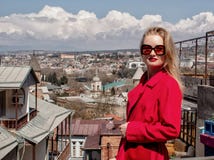 En h?rlig ung flickablondin i solglas?gon och ett r?tt lag, st?llningar p? bakgrunden av staden av Tbilisi royaltyfria bilder En h?rlig ung flickablondin i solglas?gon och ett r?tt lag, st?llningar p? bakgrunden av staden av Tbilisi royaltyfria bilder