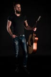 En karismatisk och stilfull man med ett skägg står hellång med en akustisk gitarr på en svart isolerad bakgrund Lodlinje fra royaltyfri fotografi En karismatisk och stilfull man med ett skägg står hellång med en akustisk gitarr på en svart isolerad bakgrund Lodlinje fra royaltyfri fotografi