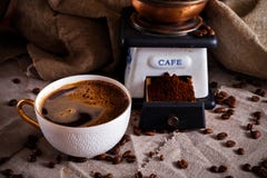 En kopp av svart kaffe, ett kaffe maler och spridda kaffebönor på en tabell som täckas med säckväv arkivfoto En kopp av svart kaffe, ett kaffe maler och spridda kaffebönor på en tabell som täckas med säckväv arkivfoto