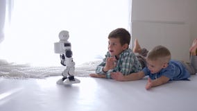 Enfants et avenir, enfants modernes mentant sur le plancher et mains de ondulation au robot de humanoïde qui va à eux Enfants et avenir, enfants modernes mentant sur le plancher et mains de ondulation au robot de humanoïde qui va à eux