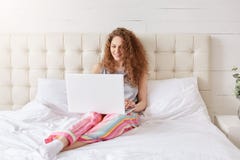 Erfüllte Schönheit arbeitet an Laptop im Bett, gekleidet in den Pyjamas, genießt den Komfort und zu Hause ist, angeschlossen an H lizenzfreies stockbild Erfüllte Schönheit arbeitet an Laptop im Bett, gekleidet in den Pyjamas, genießt den Komfort und zu Hause ist, angeschlossen an H lizenzfreies stockbild