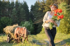 Estilo rústico, mulher feliz madura no chapéu com os ramalhetes de flores das papoilas que anda ao longo da estrada secundária imagem de stock Estilo rústico, mulher feliz madura no chapéu com os ramalhetes de flores das papoilas que anda ao longo da estrada secundária imagem de stock