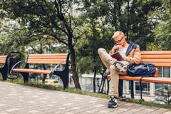 Estudante universitário com o livro de leitura da trouxa que anda no parque do outono que senta-se no banco fotografia de stock Estudante universitário com o livro de leitura da trouxa que anda no parque do outono que senta-se no banco fotografia de stock