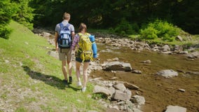 Ett ungt par av turister som promenerar en bergflod mot en bakgrund av det gröna skogloppet och aktiv lager videofilmer Ett ungt par av turister som promenerar en bergflod mot en bakgrund av det gröna skogloppet och aktiv lager videofilmer