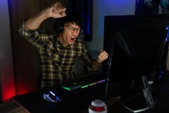 Excited Asian Man Pro Gamer Sitting At The Table, Playing And Winning In Online Video Games On A Computer And Smartphone, Royalty Free Stock Photo