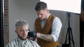 Fabbricazione del taglio di capelli Giovane uomo barbuto che ottiene taglio di capelli dal barbiere mentre sedendosi nella sedia stock footage Fabbricazione del taglio di capelli Giovane uomo barbuto che ottiene taglio di capelli dal barbiere mentre sedendosi nella sedia stock footage