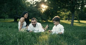Familia feliz de tres que mienten en la hierba en parque del verano Juegan y sonríen 4K Cámara lenta almacen de video Familia feliz de tres que mienten en la hierba en parque del verano Juegan y sonríen 4K Cámara lenta almacen de video