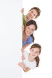 Familia feliz que mira a escondidas de la cartelera en blanco imagen de archivo Familia feliz que mira a escondidas de la cartelera en blanco imagen de archivo