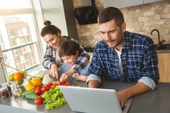 Familie die thuis in de scherpe tomaten van de keuken zich moeder met zoon verenigen terwijl vader speelspel op opgewekt laptop stock foto's Familie die thuis in de scherpe tomaten van de keuken zich moeder met zoon verenigen terwijl vader speelspel op opgewekt laptop stock foto's