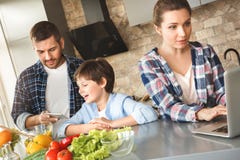 Family at home standing in kitchen together father and son playing game on digital tablet while mother working on laptop royalty free stock image Family at home standing in kitchen together father and son playing game on digital tablet while mother working on laptop royalty free stock image
