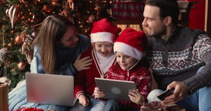 Family with kids using electronic devices sit near Christmas tree