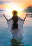 Fantasy woman standing in water hands raised to sky, praying to sun. Slovenian girl in herbal wreath on head, long hair