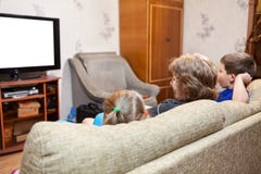Farmor med två unga ungar som hemma sitter på soffan och hållande ögonen på tv, isolerad vit skärm royaltyfri foto Farmor med två unga ungar som hemma sitter på soffan och hållande ögonen på tv, isolerad vit skärm royaltyfri foto