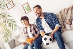 Father and little son at home sitting on sofa boy holding potato chip dad with ball watching football smiling joyful royalty free stock photo Father and little son at home sitting on sofa boy holding potato chip dad with ball watching football smiling joyful royalty free stock photo