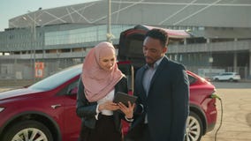 Female dealer in hijab showing something to African man in suit on digital tablet. Man and and woman standing together