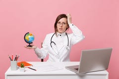 Female doctor sit at desk work on computer with medical document hold globe in hospital isolated on pastel pink wall royalty free stock photo Female doctor sit at desk work on computer with medical document hold globe in hospital isolated on pastel pink wall royalty free stock photo