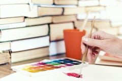Female hand draws with a brush and watercolor paint on a sheet of paper on wooden table with stack of books background
