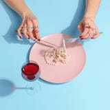 Female hands cut pearl beads with a knife and fork in a pink plate on a blue table. Creative concept. Top view royalty free stock photography Female hands cut pearl beads with a knife and fork in a pink plate on a blue table. Creative concept. Top view royalty free stock photography