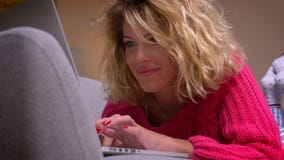Femme au foyer blonde en gros plan dans le chandail rose se trouvant sur l'estomac observant dans l'ordinateur portable smilingly clips vidéos Femme au foyer blonde en gros plan dans le chandail rose se trouvant sur l'estomac observant dans l'ordinateur portable smilingly clips vidéos