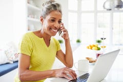 Femme à l'aide de l'ordinateur portable et parlant au téléphone dans la cuisine à la maison Images libres de droits
