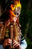 Fille avec le masque shamanic de plume et robe historique dans des environs de région boisée jouant la cannelure ornementale en b photos stock Fille avec le masque shamanic de plume et robe historique dans des environs de région boisée jouant la cannelure ornementale en b photos stock