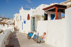 Fira town street with white houses and a white tourist with glasses sits with her back to the wall next to a motorcycle stock image Fira town street with white houses and a white tourist with glasses sits with her back to the wall next to a motorcycle stock image