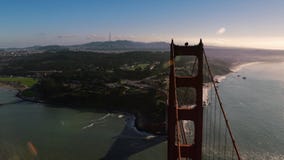 Flying over Golden Gate bridge near South tower towards San Francisco presidio, rocky shore, sunny day scenery