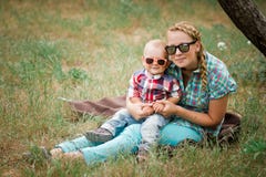 Forme o bebê nos óculos de sol que sentam-se com a mãe sob a árvore imagens de stock royalty free Forme o bebê nos óculos de sol que sentam-se com a mãe sob a árvore imagens de stock royalty free