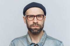 Foto do homem à moda seguro inteligente com a barba grossa escura e do bigode, olhares seriamente na câmera, poses contra o stu c imagem de stock Foto do homem à moda seguro inteligente com a barba grossa escura e do bigode, olhares seriamente na câmera, poses contra o stu c imagem de stock